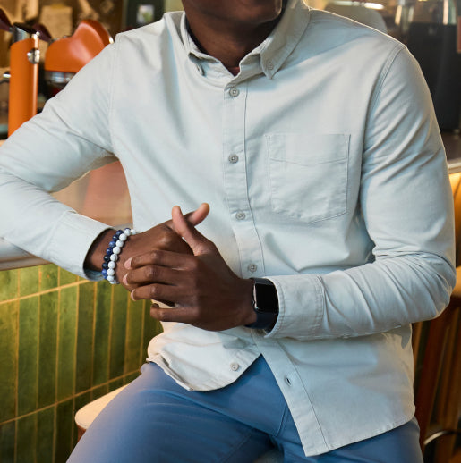 A man sitting on a bar stool, dressed in blue pants and a white collared long-sleeve shirt.